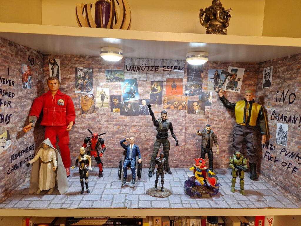 Photo of a diorama of action figures, lit by two small LED lights: The Shrine of the Useless Eaters. They stand against a brick patterned background, covered in photos of other cyborg, disabled or other Nazi-targeted peoples. Anti-fascist grafitti is on the walls; at the back is a faux-metal plaque with the German phrase “Unnütze Esser”.
